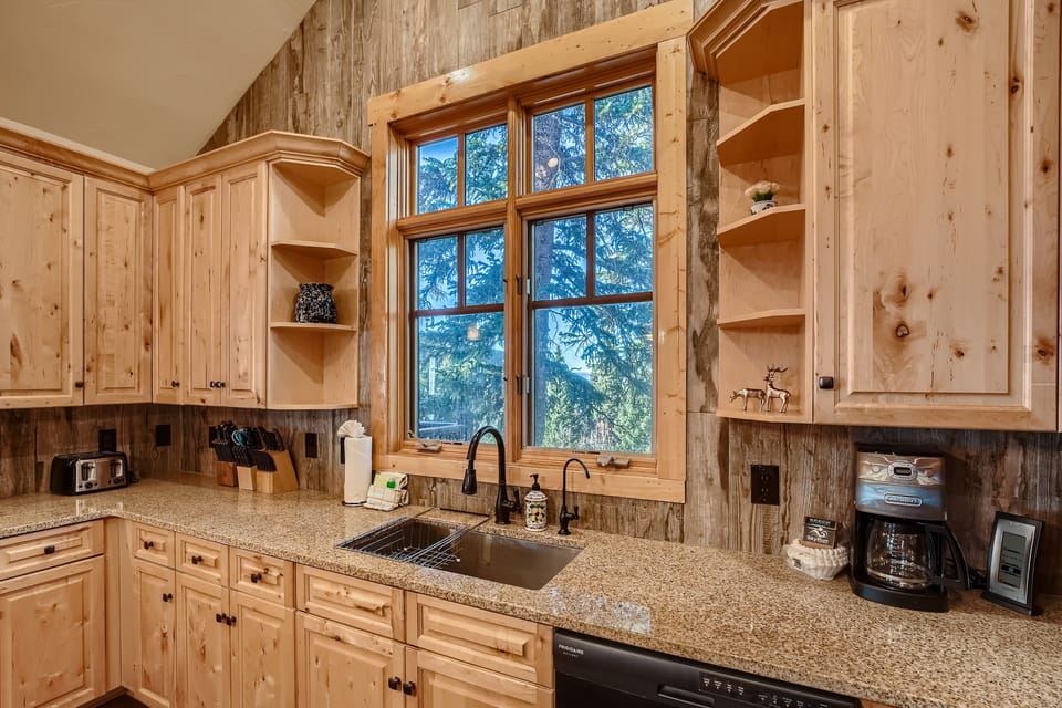 A rustic kitchen features light wood cabinets, a stone countertop, a window with a forest view, a sink with a black faucet, and various kitchen appliances and utensils.