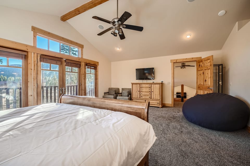 Spacious bedroom with wooden furniture, a double bed, a large bean bag, flat-screen TV, and seating area. Room has large windows and a door leading to a balcony with forest views.