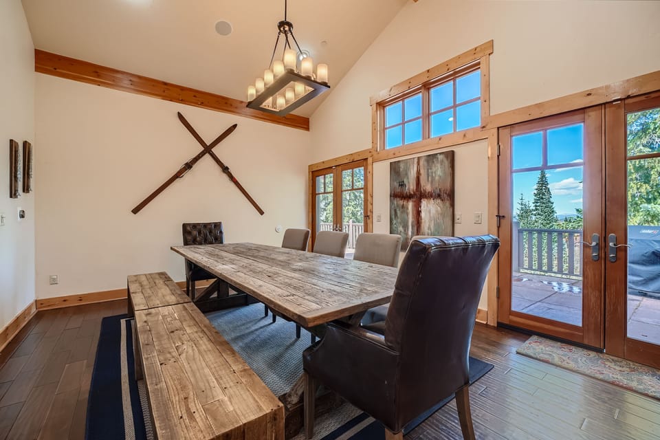 A rustic dining room with a large wooden table, leather chairs, a bench, and ski decor on the wall. The room has high ceilings, large windows, and glass doors leading to a wooden deck with forest views.