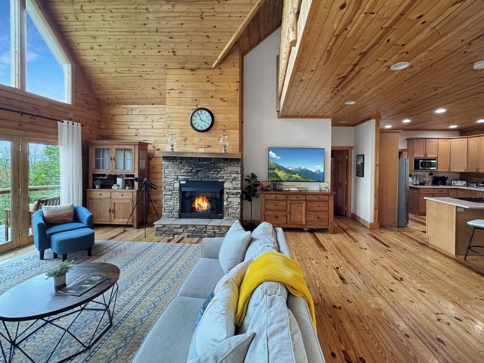 Soak in stunning mountain views from this cozy, light-filled cabin! Featuring floor-to-ceiling windows, vaulted wood ceilings, a stone fireplace, smart TV, and spacious seating—this living room is perfect for relaxing after a day of outdoor adventure.