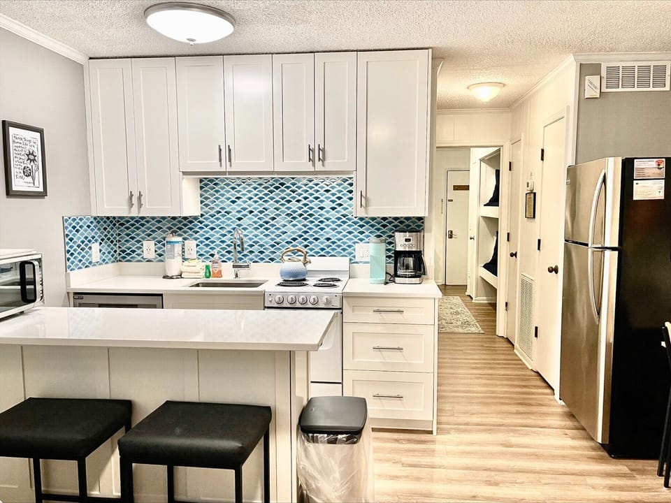 Modern kitchen with white cabinetry, blue tiled backsplash, stainless steel appliances, and a central island with two black stools. Adjacent hallway and wall art visible.