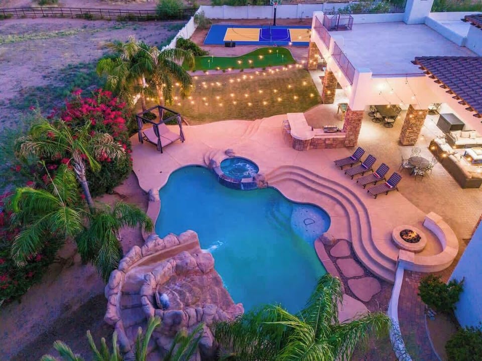 Aerial view showcasing the property's expansive outdoor amenities, including a freeform pool with adjacent hot tub, basketball court, covered patio with fire pit, and festive string lighting throughout the landscaped grounds.