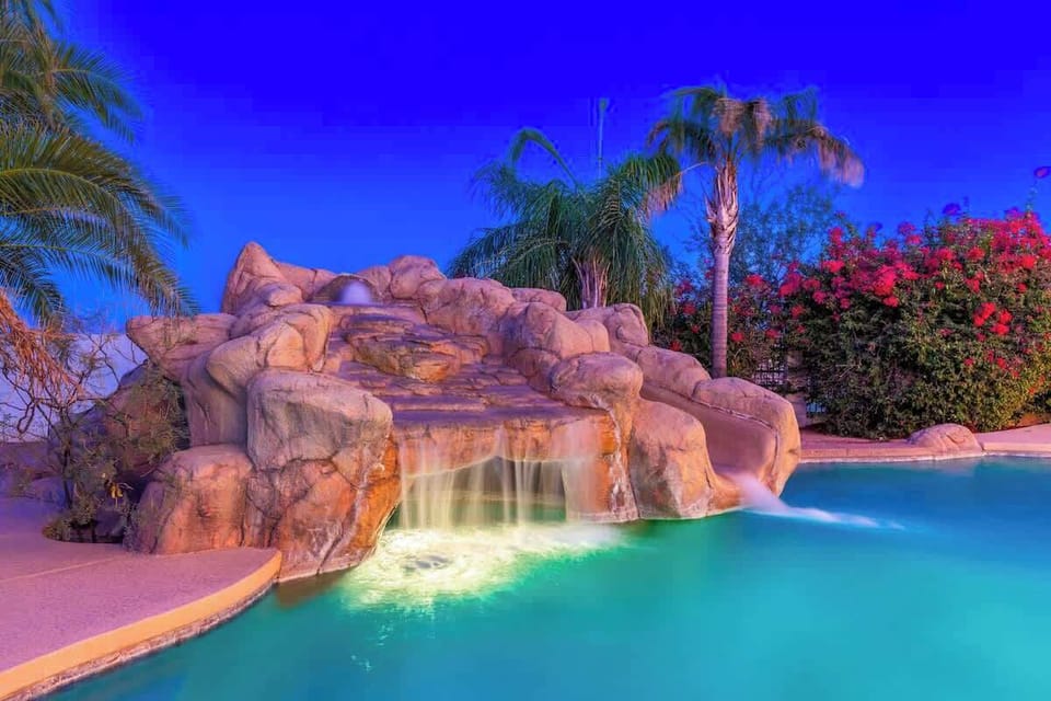 A tropical paradise awaits with an illuminated waterfall cascading into a pristine pool, surrounded by palm trees and vibrant blooms under a stunning twilight sky.