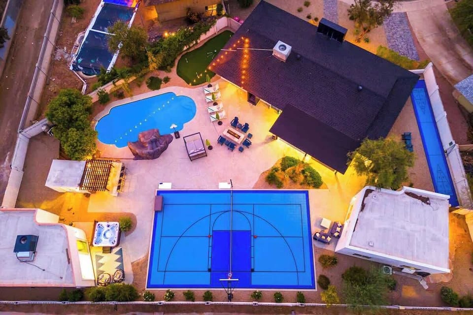 Aerial view showcasing a vacation rental property with diverse outdoor amenities including a swimming pool, sport court, hot tub, putting green, and covered patio area surrounded by mature landscaping and neighboring residences.