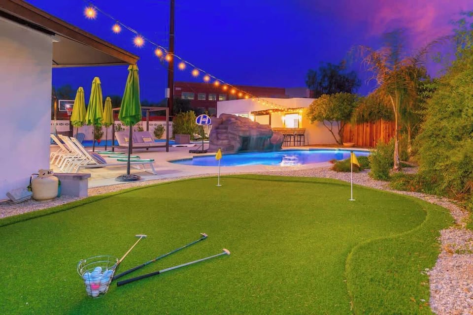 The property features a resort-style backyard with a mini golf green, cascading waterfall pool, spa, lounge seating under umbrellas, and festive string lights—all set against a stunning evening sky.