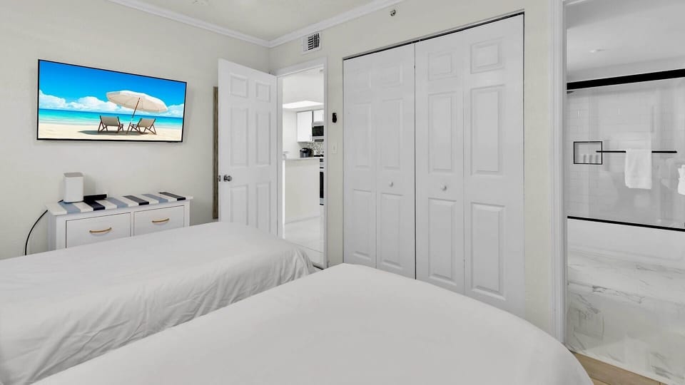 Your bright bedroom features two plush beds, a wall-mounted TV, ample wardrobe space, and a modern en-suite bathroom—perfect for resting after beach adventures.