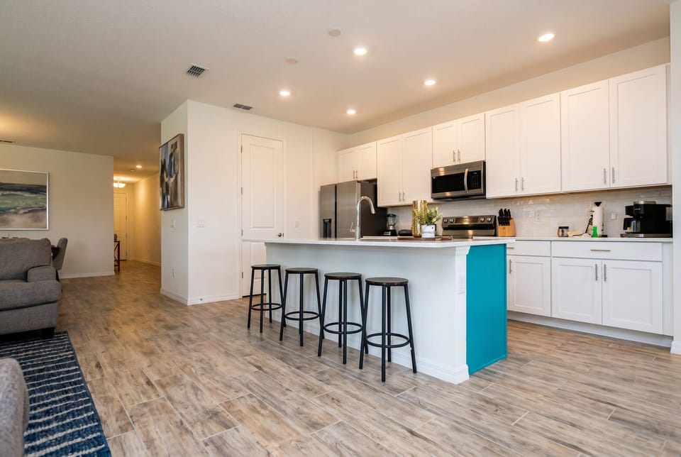 Welcome to our bright, airy kitchen with modern white cabinetry and a soft blue island, perfect for meals and casual chats. The warm wood-like flooring enhances the space, inviting abundant natural light for a cozy atmosphere.