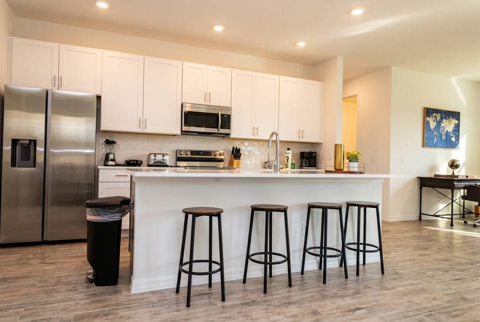 The sleek kitchen boasts a spacious island with modern bar stools for casual dining or morning coffee. Bright white cabinetry and stainless steel appliances create a clean, functional space ready for your culinary adventures.