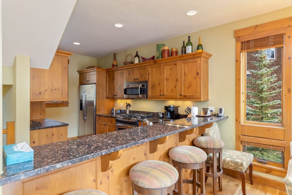 A wooden kitchen with a granite countertop, bar stools, stainless steel appliances, and various items on the counters and shelves. A window on the right offers a partial view of outdoors.