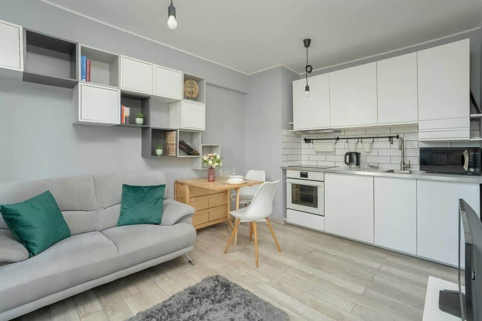 An open living space with a gray sofa, teal pillows, white kitchen cabinets, and a small wooden dining table.

