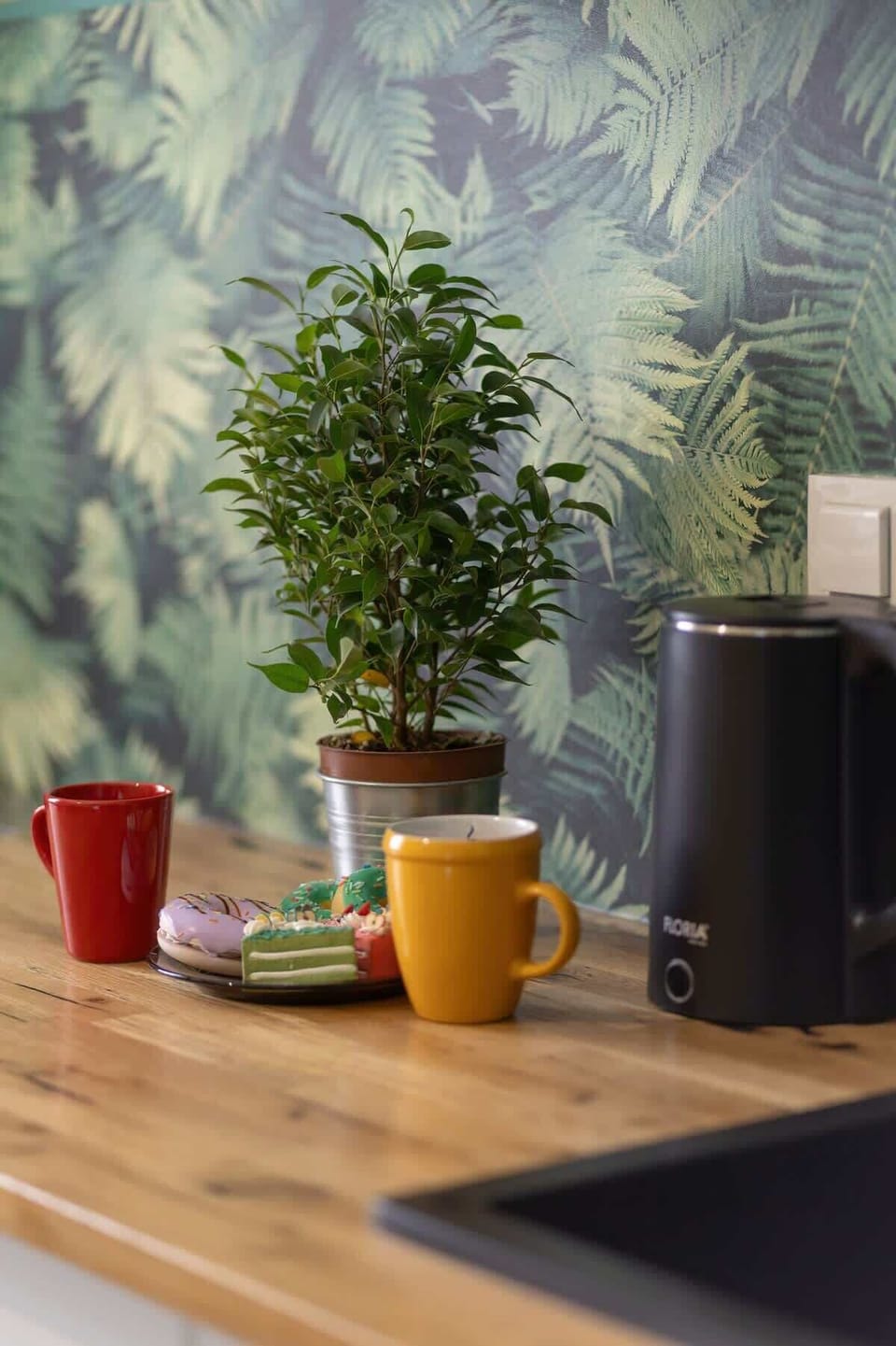 A kitchen counter with a wooden surface, a potted plant, and colorful coffee mugs, set against a tropical-themed backsplash.
