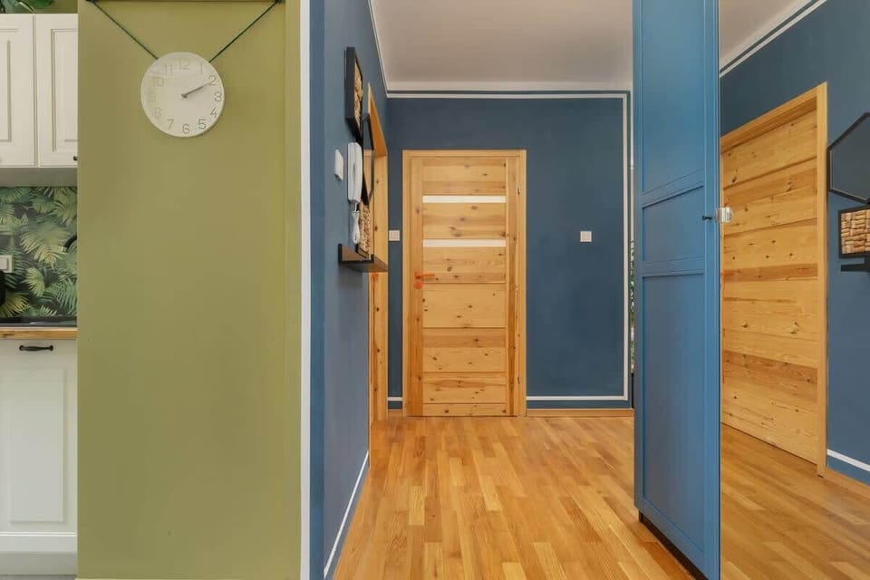 A hallway with blue walls, wooden doors, and a modern clock, leading to different rooms in the apartment.
