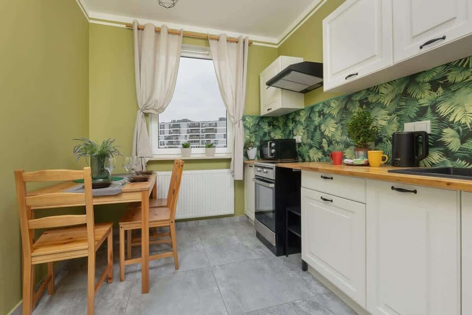A compact kitchen with green walls, white cabinets, and a small dining table near the window.
