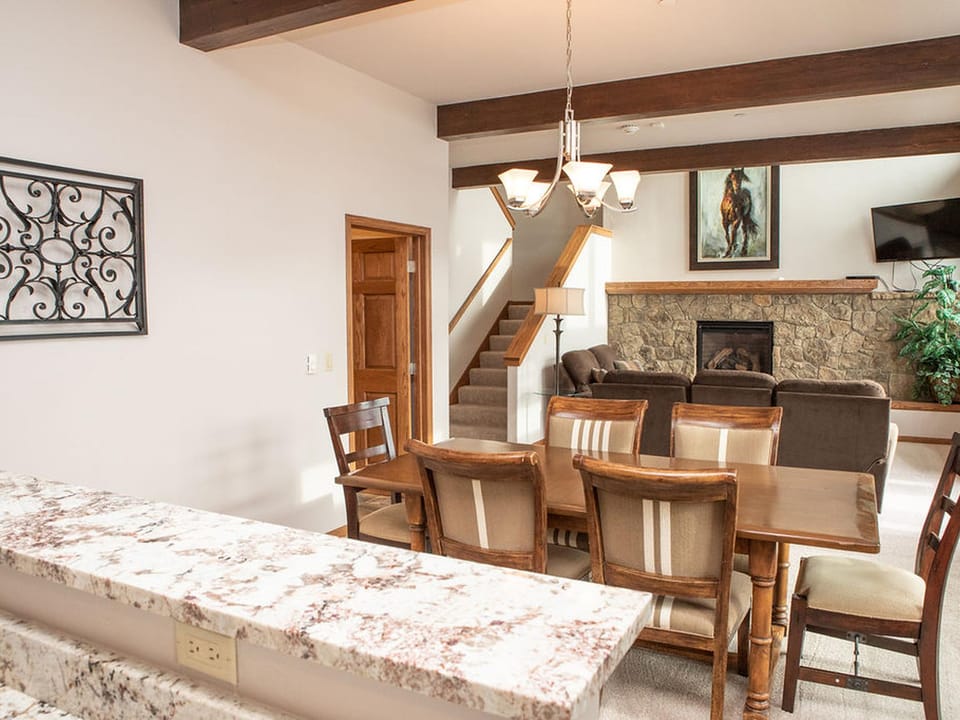 A living and dining area with wooden furniture, a stone fireplace, and a decorative wall hanging. A staircase leads upstairs, and a painting and TV are mounted on the walls.