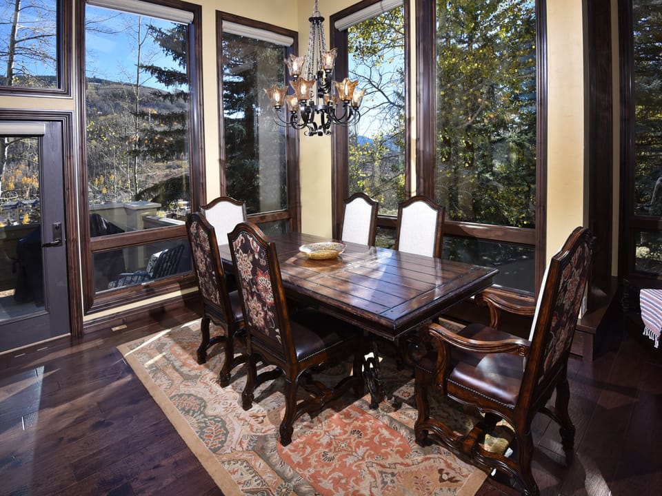 A dining room with a wooden table and six chairs, surrounded by large windows. A chandelier hangs above the table, and a patterned rug lies underneath. Trees and outside scenery are visible through the windows.