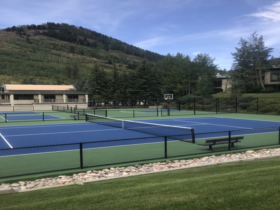 Outdoor tennis and pickleball courts with a basketball hoop in the background, surrounded by trees and mountains under a blue sky.
