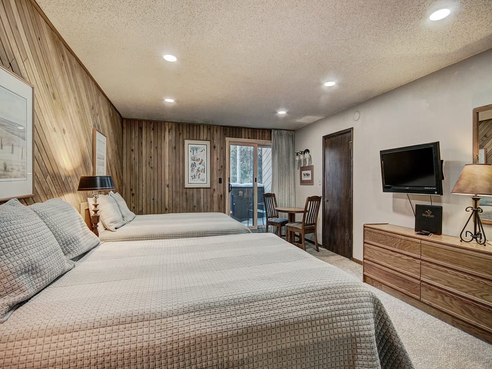 A cozy bedroom with two beds, a wooden nightstand, a TV mounted on the wall, and a small table with two chairs. The walls are partially covered with wood paneling.