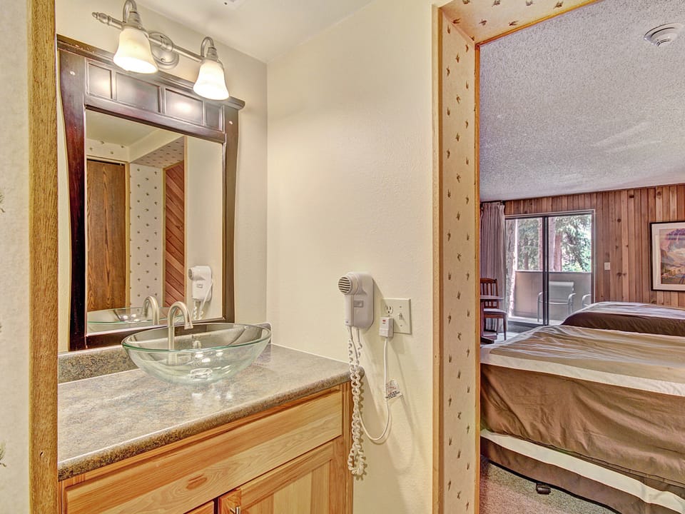 A hotel room with a view of the main bedroom featuring two beds. In the foreground, there is a small vanity area with a glass sink, mirror, and a wall-mounted hairdryer. The room has wooden accents.