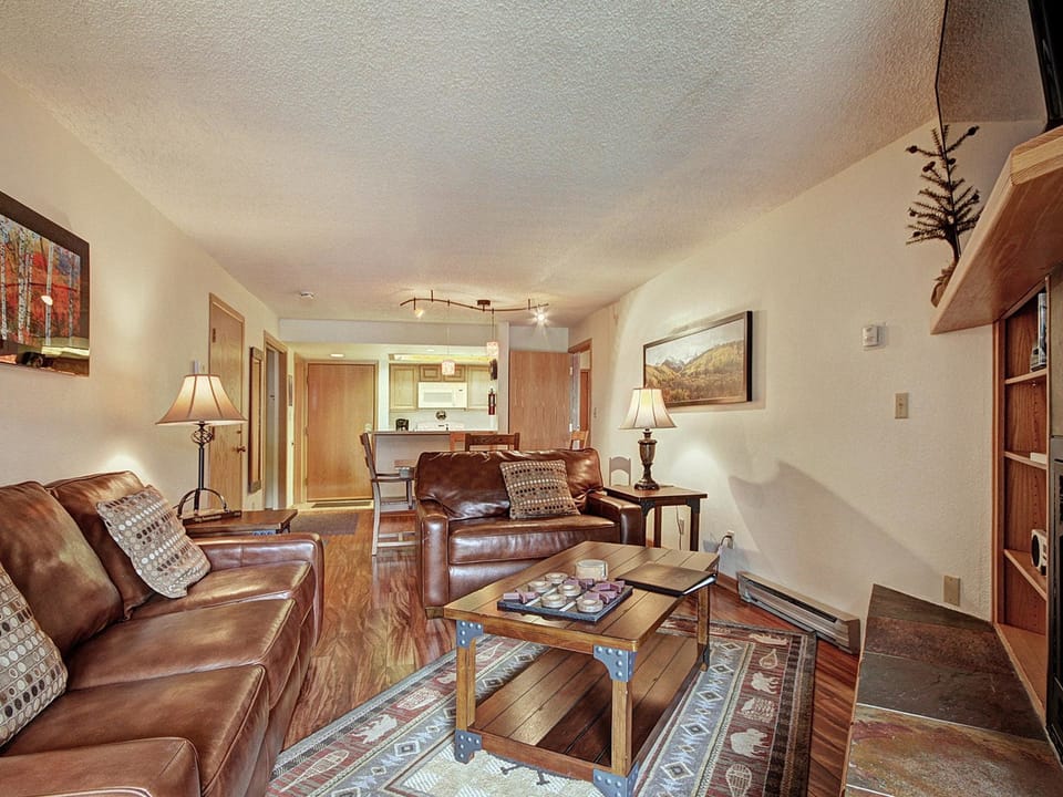 A cozy living room with leather sofas, a coffee table, and lamps, leading into a kitchen area. A painting and a framed picture adorn the walls.