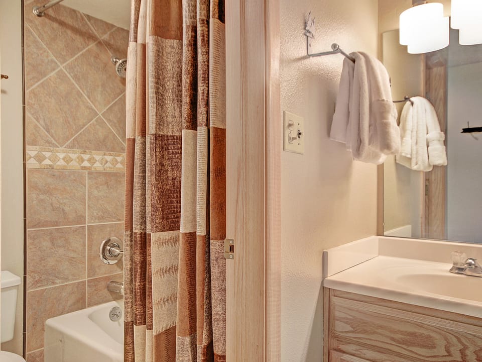 Bathroom with a toilet, a bathtub with tiled walls, a shower curtain, a vanity with a sink, and a mirror above. Two white towels hang on a wall-mounted towel rack.