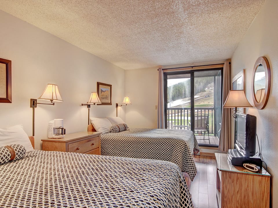 A hotel room with two double beds, bedside lamps, framed pictures, a TV, dresser, and sliding glass door leading to a balcony with an outdoor view.