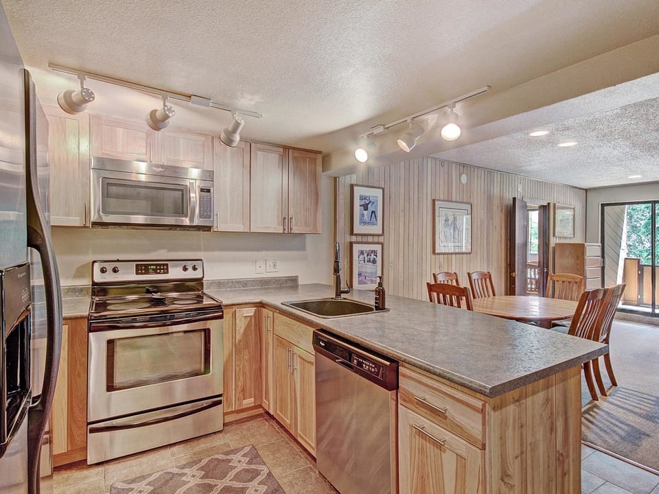 A modern kitchen with stainless steel appliances, light wood cabinets, and a granite countertop island. Adjoining dining area with a wooden table and chairs, and a view of a balcony with outdoor greenery.