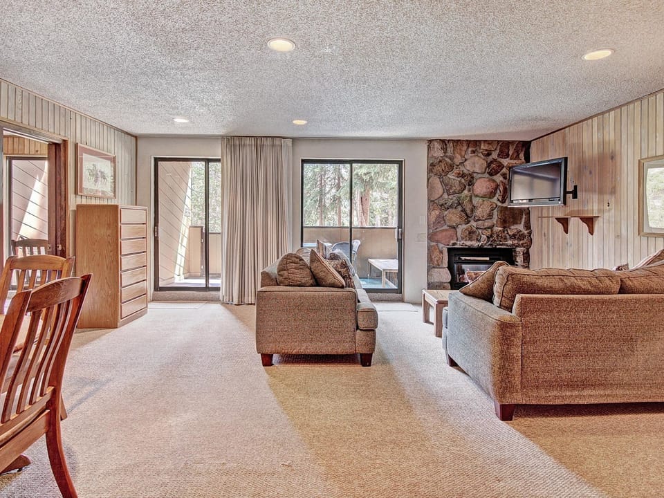 A living room features two sofas, a wall-mounted TV, a stone fireplace, and a glass sliding door leading to a balcony. The room has wood-paneled walls and a dining table with chairs on one side.