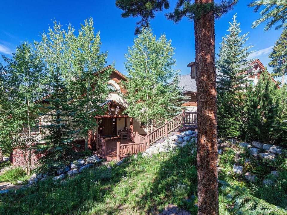 A house surrounded by tall trees and shrubs, with a wooden staircase leading to the front entrance and a sloped path in a landscaped, forested area.