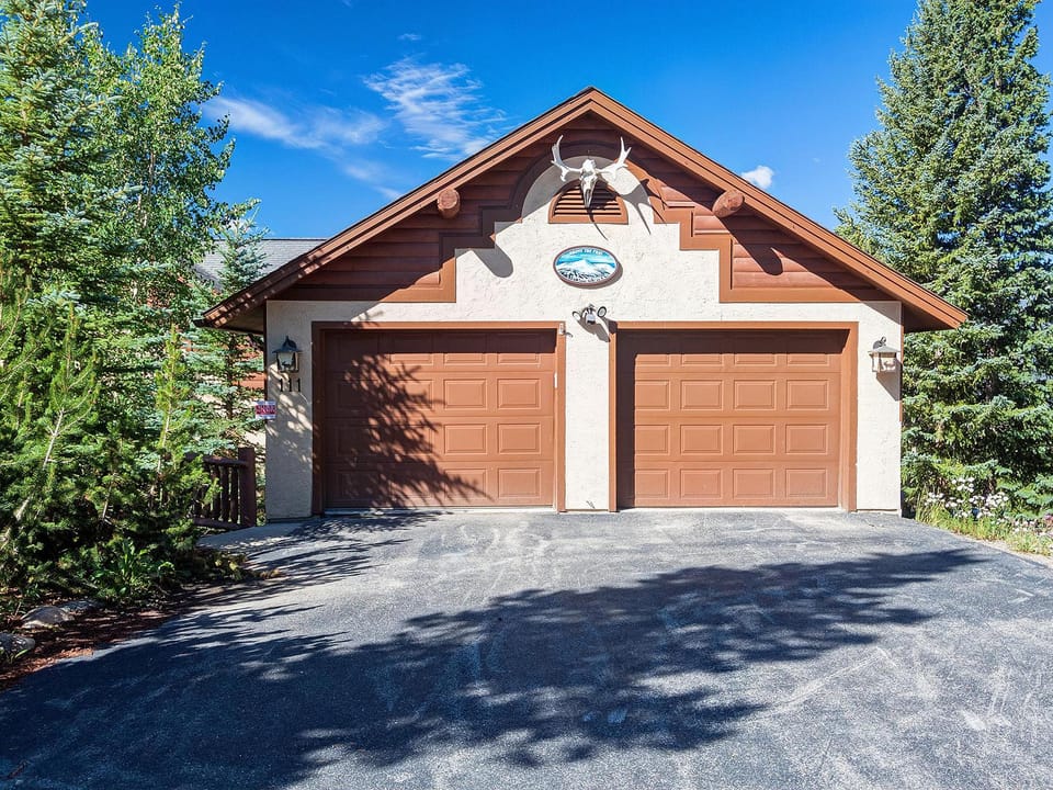 A two-door garage with a peaked roof and wall-mounted antlers above the doors, surrounded by trees and shrubs under a clear blue sky.