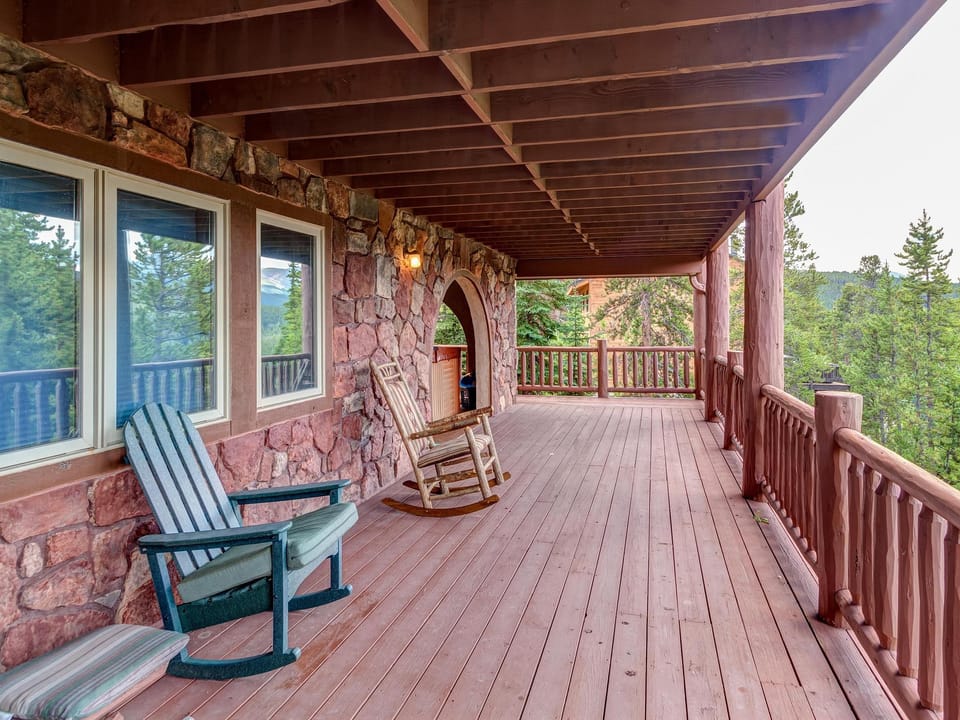A wooden porch with two chairs and a rocking chair offers a view of surrounding trees and mountains. The porch is attached to a stone exterior building with large windows.