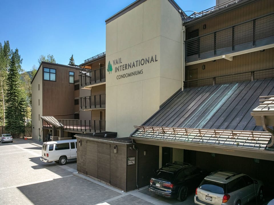 Exterior view of Vail International Condominiums, featuring multi-story buildings, parked vehicles, and surrounding trees on a sunny day.