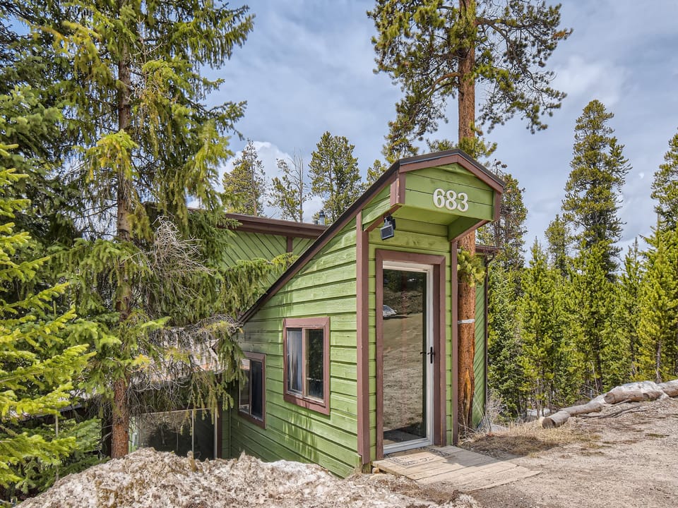 A green cabin with the number 683 on the front, surrounded by pine trees and snow on the ground. A glass door is at the entrance.