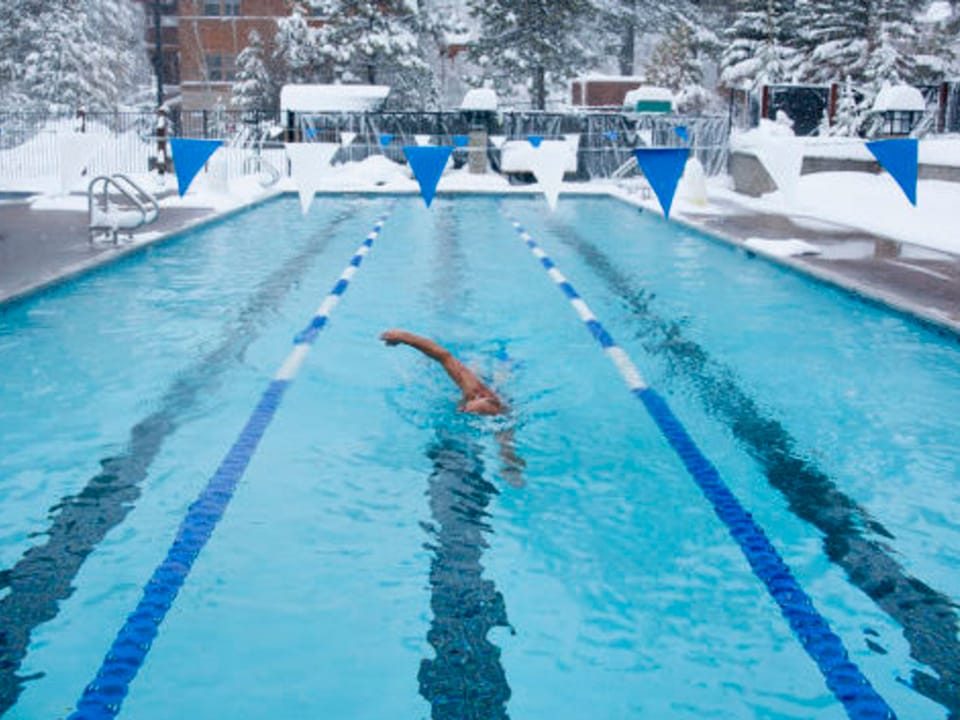 A person swimming in an outdoor pool with lane markers, surrounded by snow-covered ground and trees.