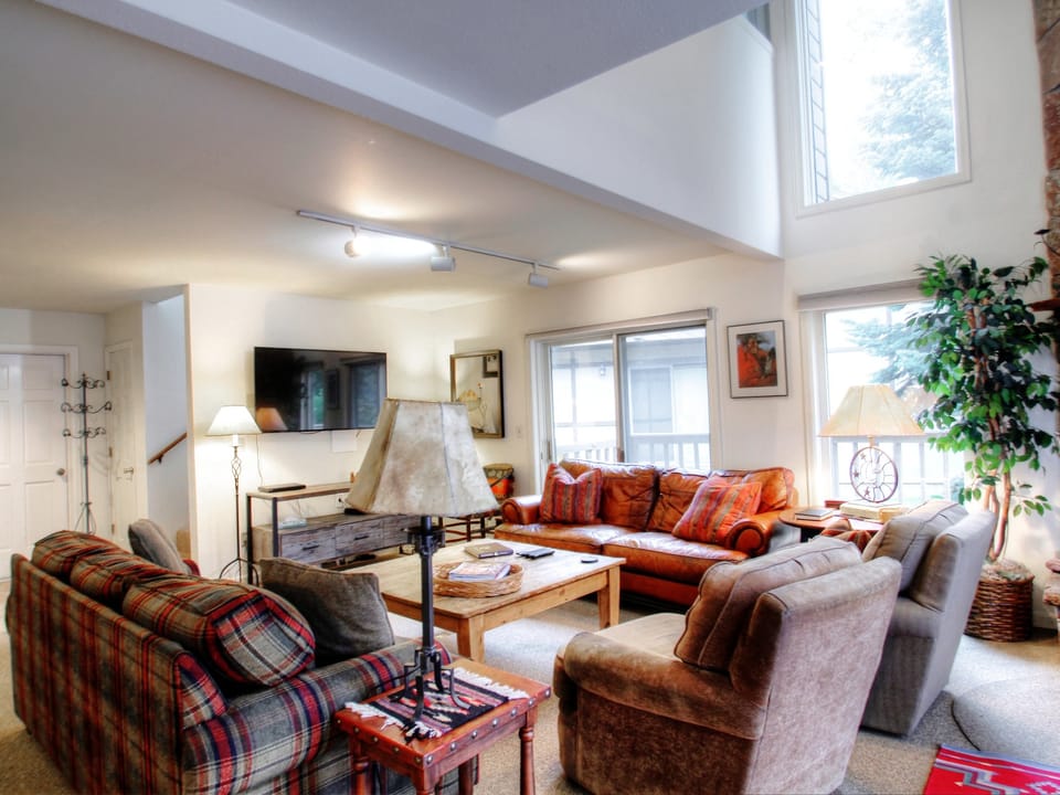 A cozy living room with various seating options, including a red couch, plaid sofa, and beige armchairs. The room has a fireplace, large windows, a wall-mounted TV, a small wooden table, and lamps.