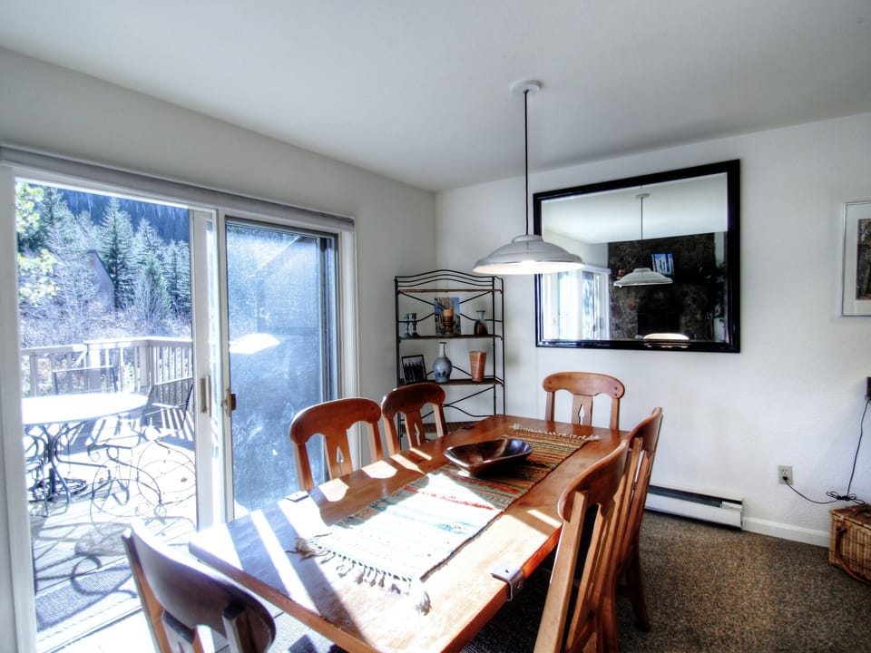 A dining room with a wooden table and chairs, pendant light, large wall mirror, and a sliding glass door leading to an outdoor deck with patio furniture.