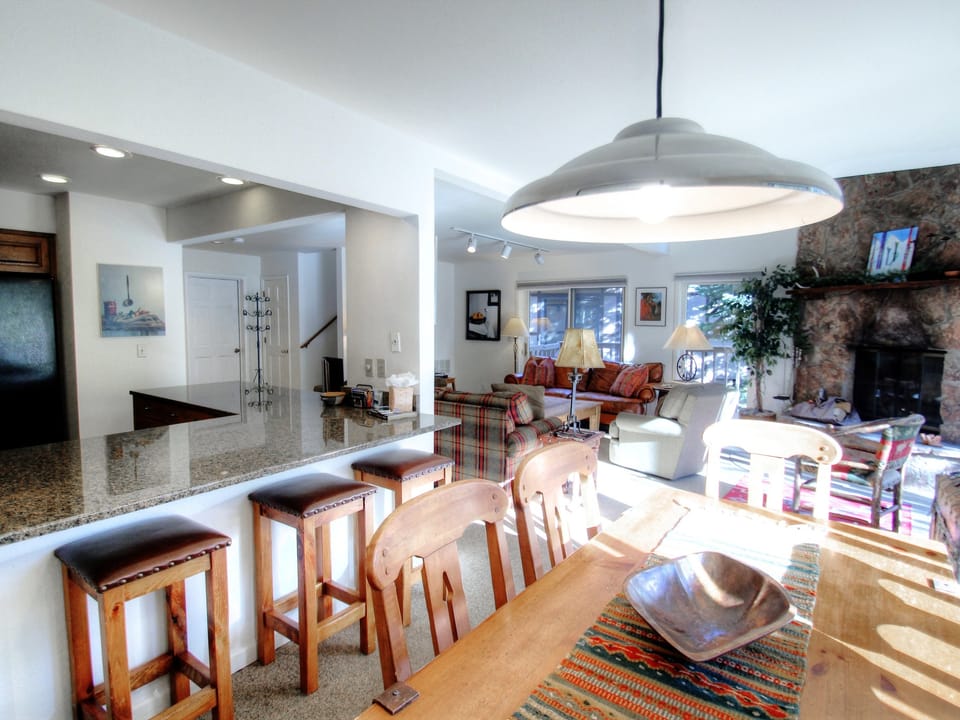 A spacious open-concept living area with a kitchen counter featuring wooden bar stools, a dining table with a pendant lamp, and a cozy living room with a stone fireplace and large windows.