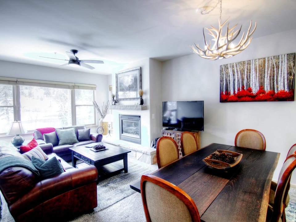 A modern living room with a leather sofa, a fireplace, a large window, a TV on a stand, and a dining table with six chairs. Antler chandelier and forest artwork enhance the decor.