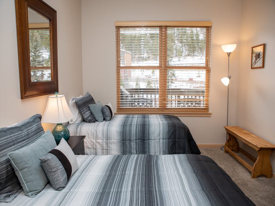 A bedroom with two twin beds featuring gray and white striped bedding, a window with blinds, a wooden bench, a framed picture on the wall, a floor lamp, and a mirror above one bed.