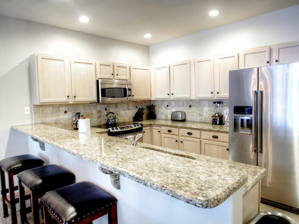 A modern kitchen with granite countertops, stainless steel appliances, light wood cabinets, and three black leather stools at the bar. The kitchen is well-lit with recessed lighting.