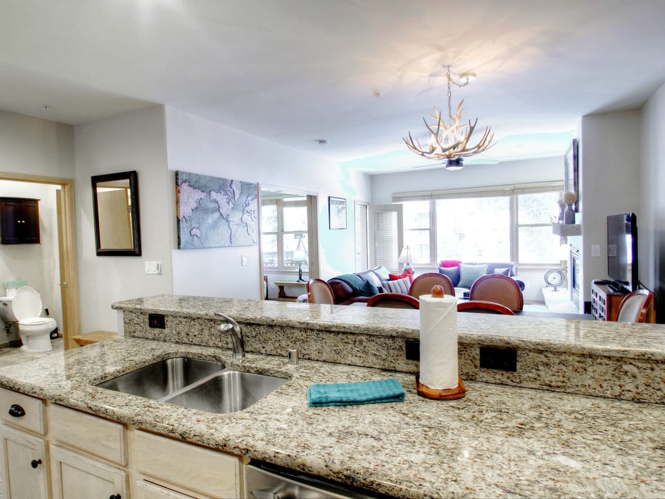 Open-concept living area with granite countertop and sink in the foreground, a living room with a couch and TV, and large windows in the background. A bathroom is partially visible on the left.