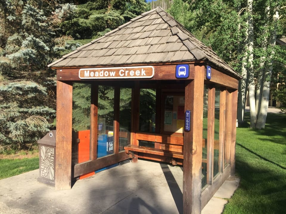 A small wooden bus shelter labeled "Meadow Creek" with bench seating and surrounded by trees is shown. A trash can and informational sign are also visible.