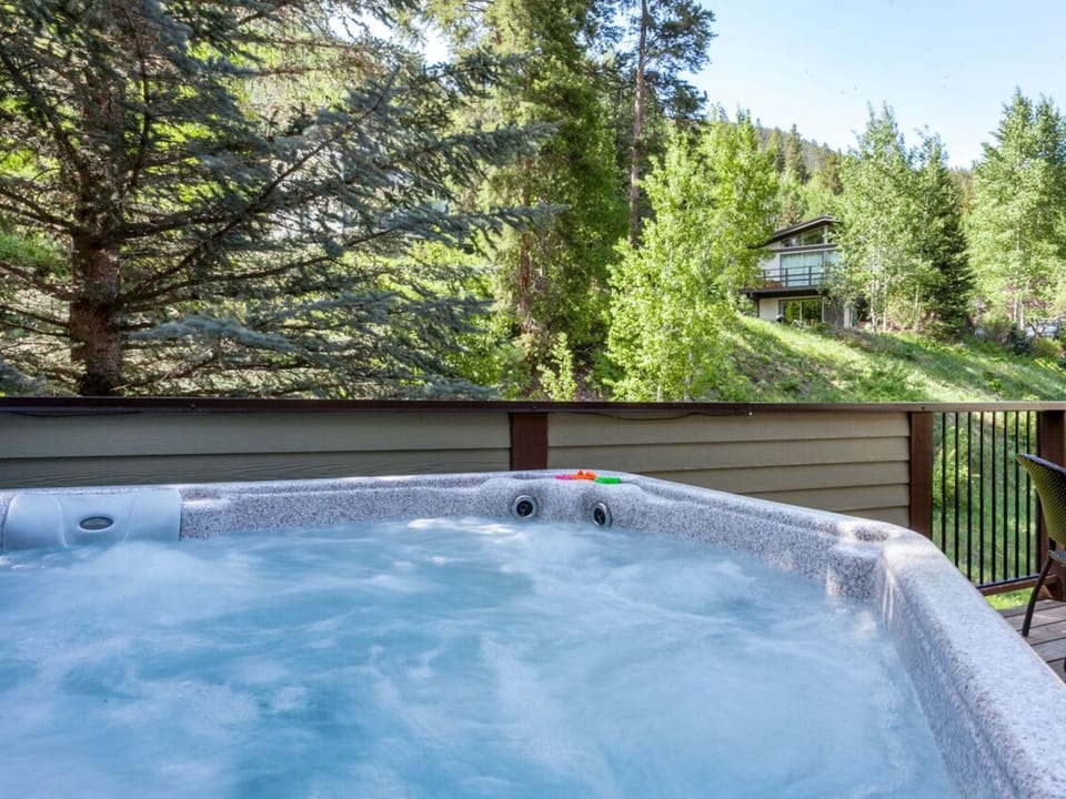 Hot tub on a wooden deck overlooking a lush, forested hillside with trees and a house in the background.