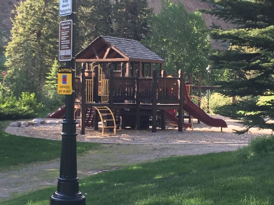 A wooden play structure with slides and climbing features is set in a forested park area. A sign near the structure warns that surfaces may be hot.