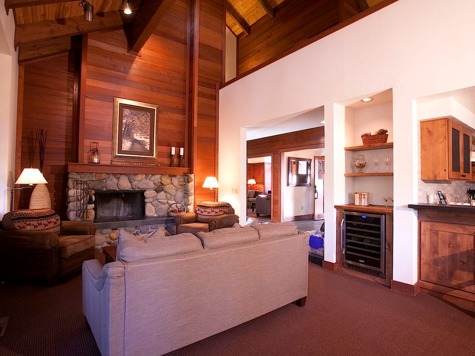 A cozy living room with a stone fireplace, wooden walls, two chairs, a sofa, and a small kitchen area with a wine cooler. Warm lighting and artwork enhance the ambiance.