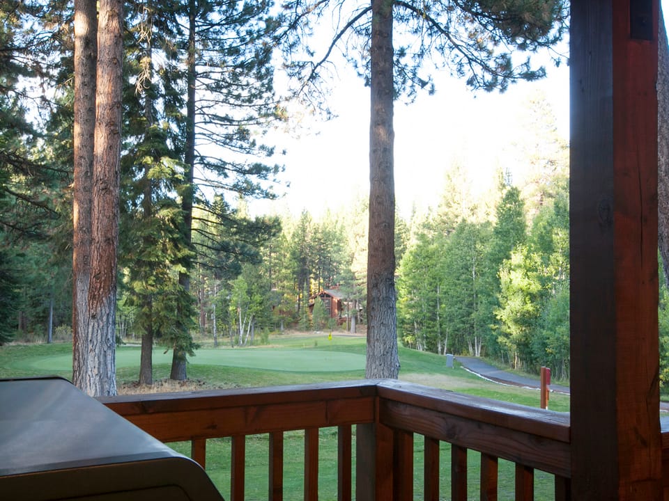 A wooden deck overlooks a lush, green golf course surrounded by tall trees. A grill is partially visible in the foreground.