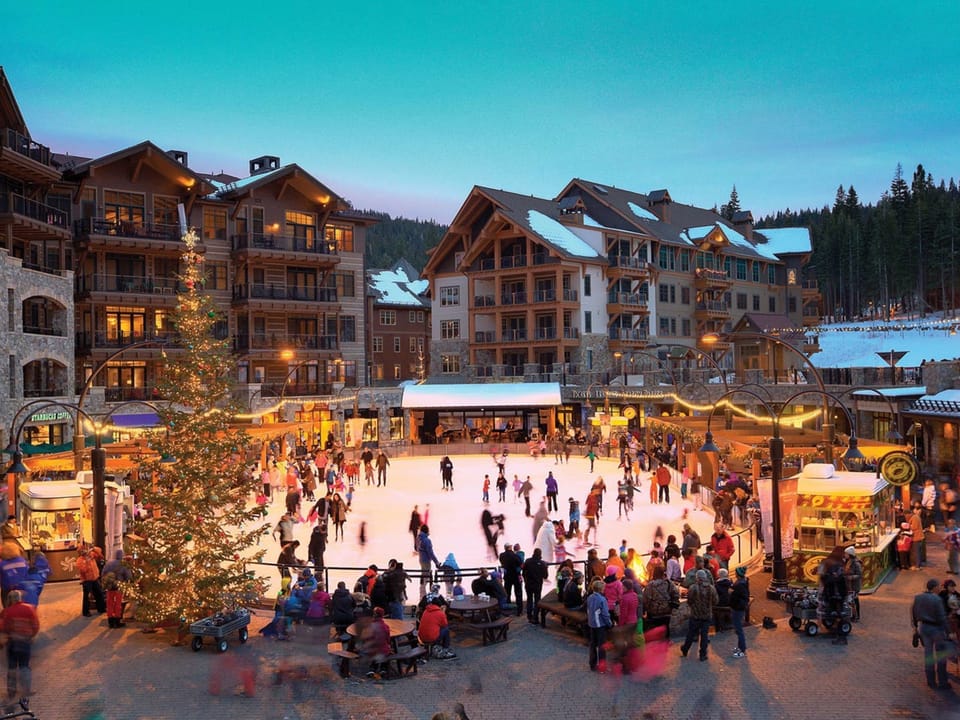 People ice skating at an outdoor rink surrounded by lit-up buildings and pine trees in the evening, with a Christmas tree nearby.