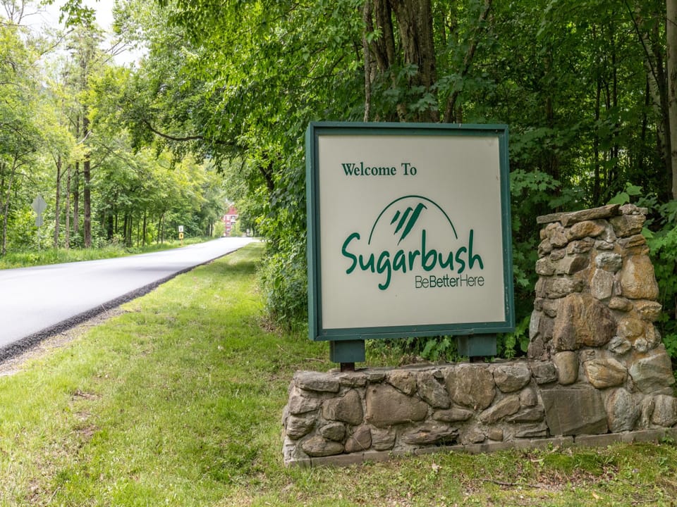 A stone base sign reads "Welcome to Sugarbush: Be Better Here," positioned beside a road and surrounded by lush green trees.