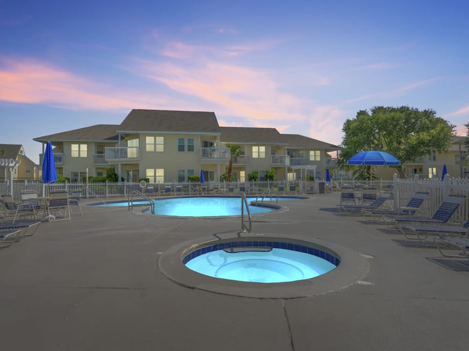 A tranquil vacation resort showcasing illuminated swimming pool and hot tub at dusk, with comfortable lounge seating and tropical palms framing the two-story accommodations.