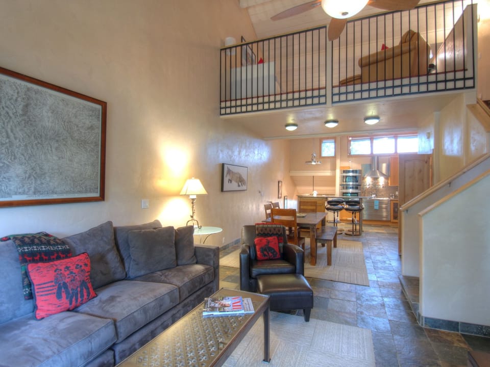 A modern living room with a gray couch, armchair, coffee table, and wall artwork. A loft area is seen above, and a dining table and kitchen are in the background. Stairs lead to the loft.