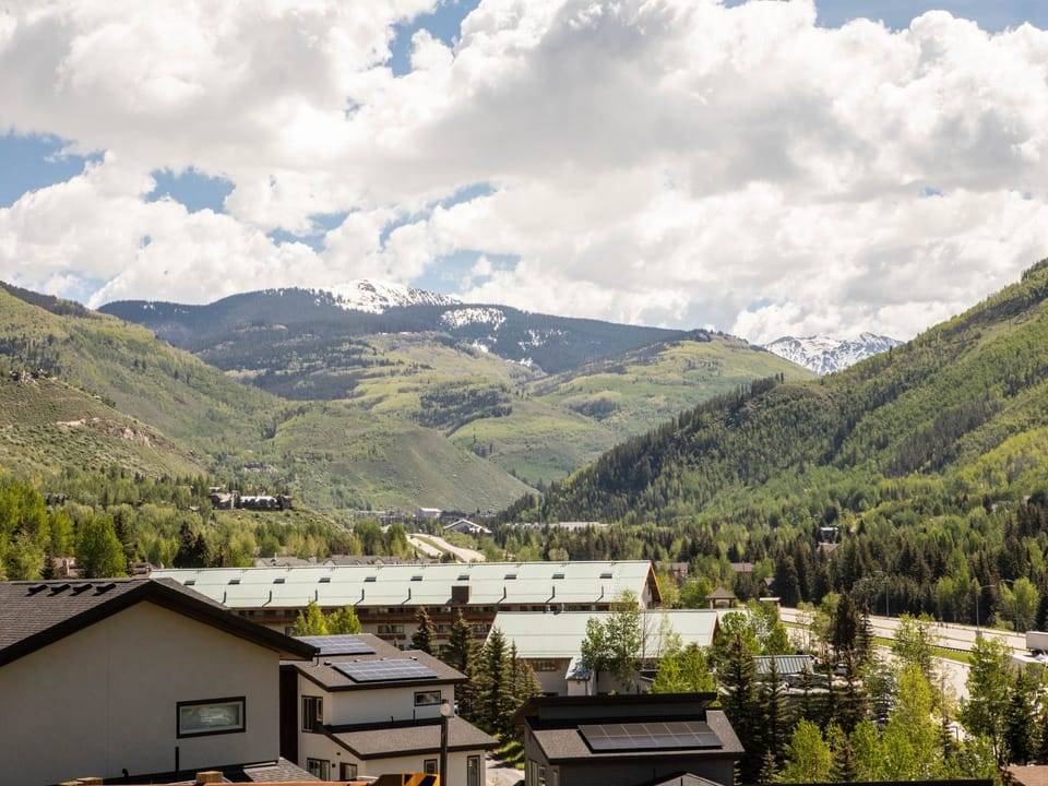A scenic view of a valley with green hills and patches of snow in the distance. Residential buildings with solar panels are visible in the foreground. The sky is partly cloudy.